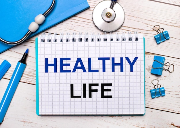 On a light wooden background, a stethoscope, a blue notepad,\
blue paper clips, a blue marker and a sheet of paper with the text\
healthy life. medical concept