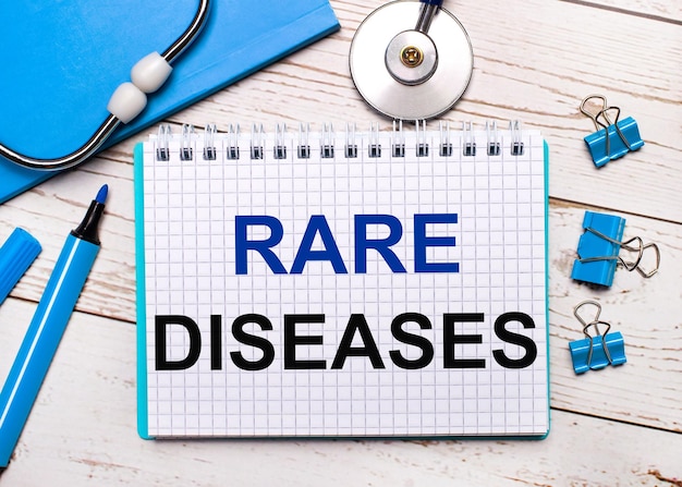 밝은 나무 배경에 청진기, 파란색 메모장, 파란색 종이 클립, 파란색 마커 및 RARE DISEASES라는 텍스트가 있는 종이 한 장. 의료 개념