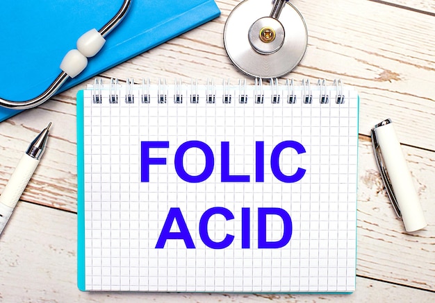 On a light wooden background there is a stethoscope, a blue notebook, a white pen and a sheet of paper with the text FOLIC ACID. Medical concept