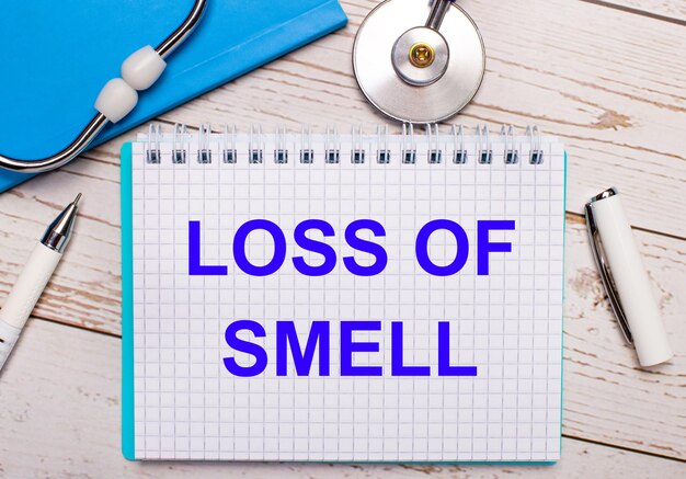 明るい木の背景には、聴診器、青いノート、白いペン、そして「LOSSOFSMELL」というテキストが書かれた一枚の紙があります。医療コンセプト