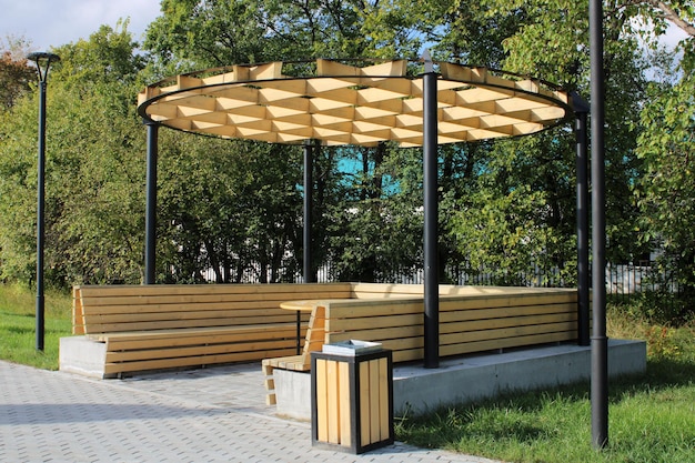 A light wooden gazebo with a bench a table and an unusual hutch in a city park Urban architecture