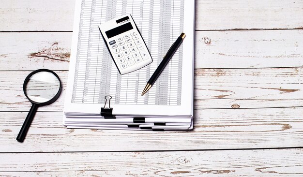 On a light wooden surface, a stack of documents, a magnifier, a pen and a white calculator