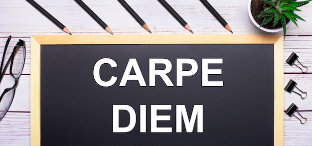 明るい木製のテーブルの上に、「CARPE DIEM」というテキストが書かれたボード、鉛筆、植物、グラス、ペーパー クリップ ビジネス コンセプト