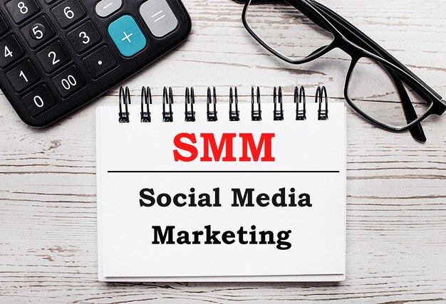 On a light wooden table calculator glasses and a blank notepad with the text SMM Social Media Marketing Business concept