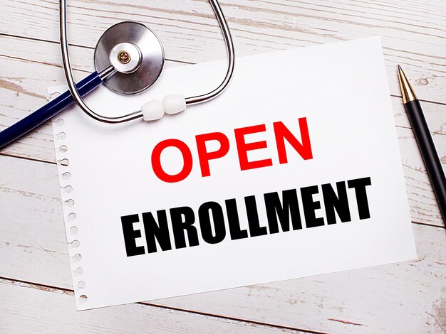 On a light wooden table there is a stethoscope a pen and a sheet of paper with the text OPEN ENROLLMENT Medical concept