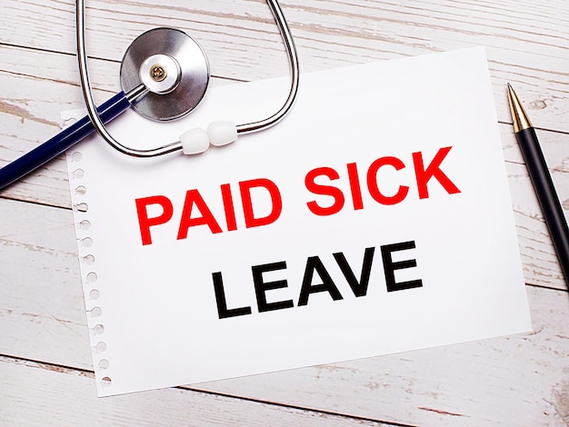 가벼운 나무 테이블에는 PAID SICK LEAVE Medical 개념이 있는 청진기, 펜, 종이 한 장이 있습니다.