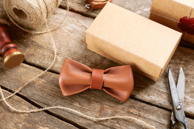 Светло-коричневый кожаный галстук-бабочка и картонная подарочная коробка на деревянном столе крупным планом