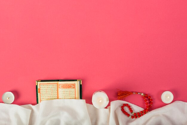 Photo lighted candle; islamic kuran; prayer beads with white clothes against red backdrop