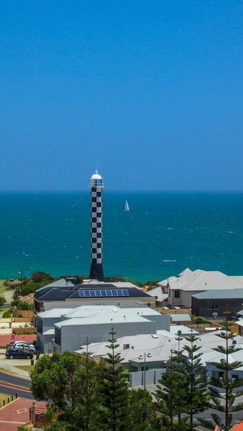 Lighthouse by sea against buildings in city against clear blue sky