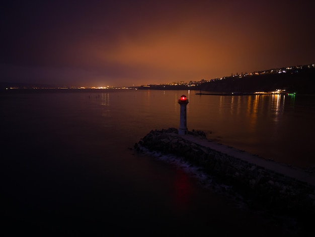 Lighthouse on the seashore with red light at night against the background of the lights of the ships