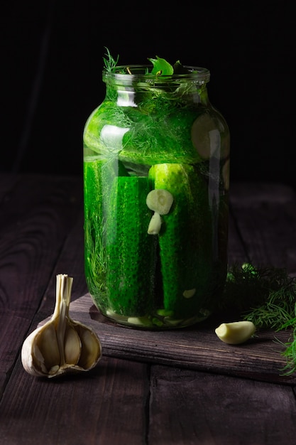 Lightly salted cucumbers in a glass jar with garlic on a dark wooden table.