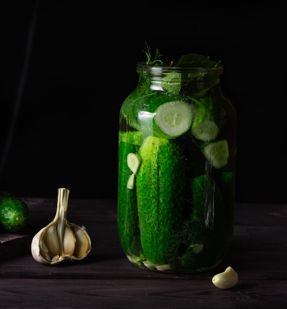 Lightly salted cucumbers in a glass jar with garlic on a dark wooden table.