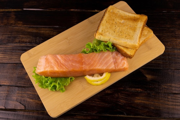 Lightly salted salmon steak with herbs and toast on a cutting board on a dark wooden background.