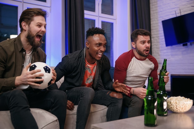 Likable joyful multiracial young guys cheering their favourite football team with screams and hands up during watching sport game on tv