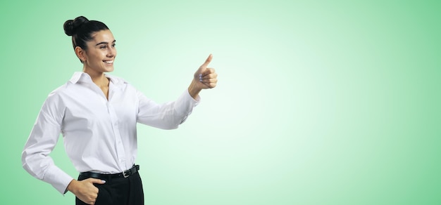 Like concept with happy young businesswoman in white shirt holding thumbs up on light green wall background with blank space for you logo mockup