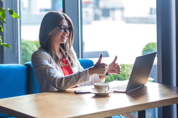 Like it Portrait of happy satisfied beautiful stylish brunette young woman in glasses sitting looking at her laptop screen toothy smile and thumbs up indoor studio shot cafe office background