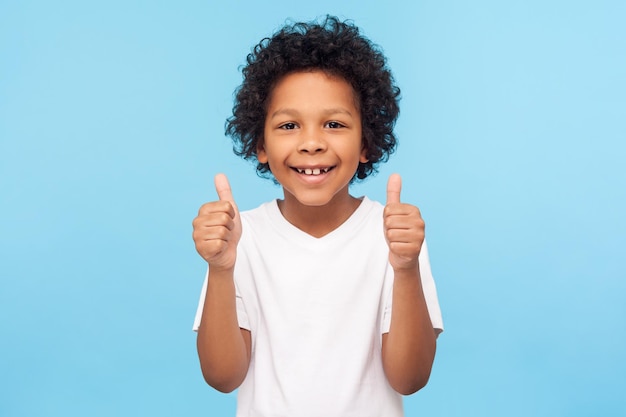 Like Portrait of happy little boy with curly hair in white Tshirt smiling at camera and doing thumbs up gesture showing agree cool approval sign indoor studio shot isolated on blue background