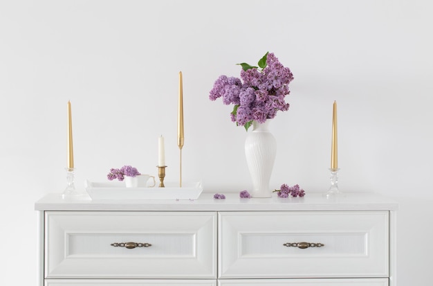Lilac flowers in white vase and golden candles on background white wall