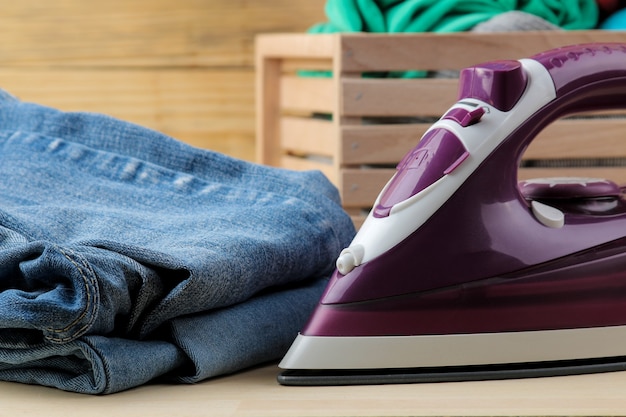 Lilac iron and a stack of clothes in a box on a natural wooden background. ironing clothes. household electrical appliances.
