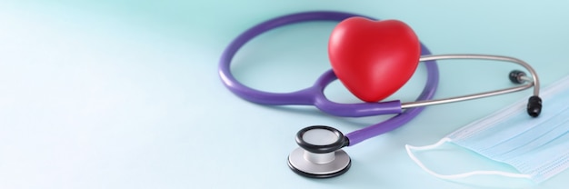 Lilac stethoscope and red toy heart with protective medical mask closeup cardiac problems