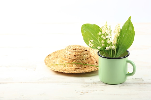 Lilies of the valley in an enamel mug with a straw hat on a white wooden background