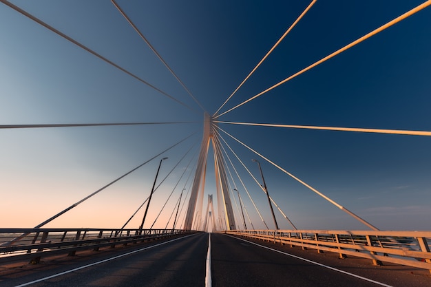 Linear perspective view of a white cable-stayed suspension bridge in the golden light of the rising sun shot from a ground level in horizontal (landscape) format