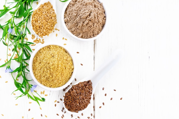 Linen bran and flour in two bowls, seeds in spoons, flax leaves and flowers on the background of a wooden board from above