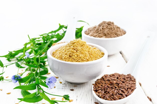 Linen bran and flour in two bowls, seeds in spoons, flax leaves and flowers on the background of a wooden board