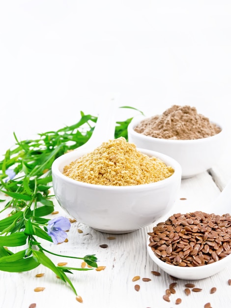 Linen bran and flour in two white bowls, seeds in spoons, leaves and blue flowers of flax on the background of a wooden board