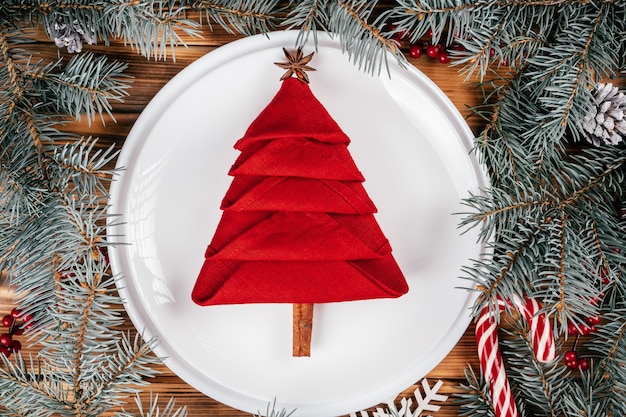 トウヒの枝の間の白い丸いプレートにクリスマスツリーの形に折りたたまれたリネンナプキン。新年のテーブル設定。上面図