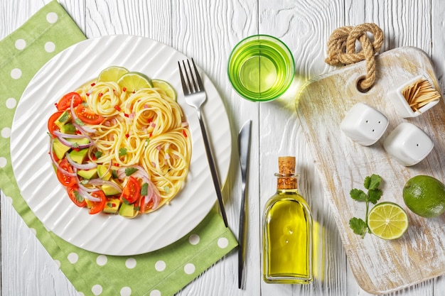 Linguine with avocado, tomatoes and lime sprinkled with chopped chili and red onion on a white plate on a wooden table, horizontal view from above, flat lay