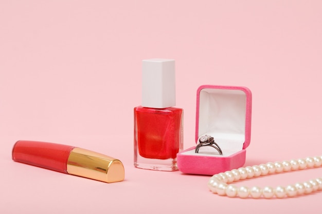 Lippenstift, nagellak, doos met zilveren ring en kralen op de roze achtergrond. Vrouwen sieraden, cosmetica en accessoires.