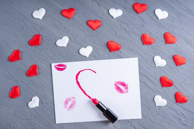 Lipstick draws a half heart shape on white paper with lipstick kisses on a gray wooden background