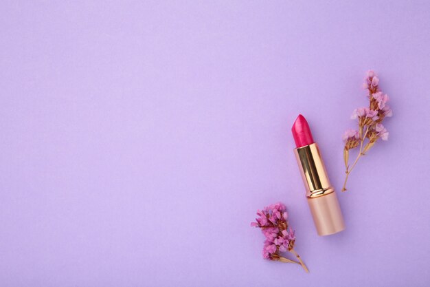 Lipstick with beautiful flowers on purple background with copy space. top view