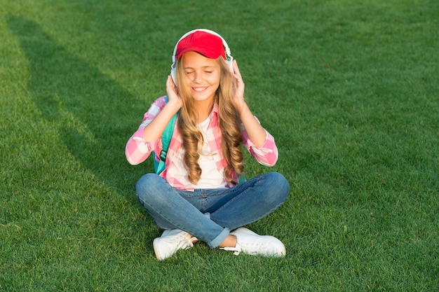 Listen music while relaxing outdoors Kid girl enjoy music green grass meadow Pleasant time Child headphones listen music Girl headphones listening music Educational podcast Enjoy every moment