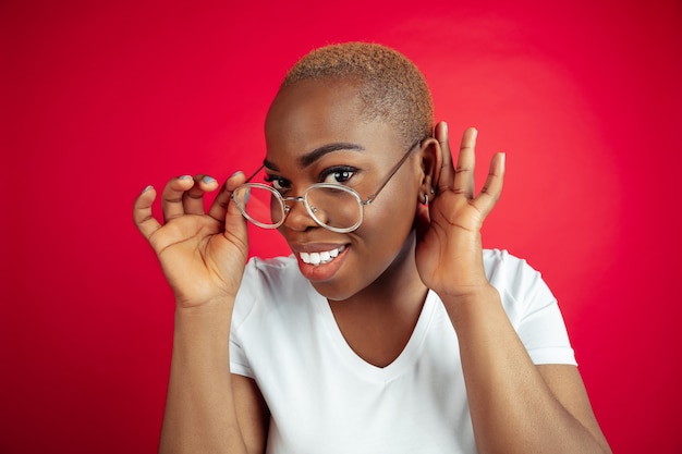 Listen secret. African-american young woman's portrait on red background. Beautiful female model in shirt. Concept of human emotions, facial expression, sales, ad, inclusion, diversity. Copyspace.