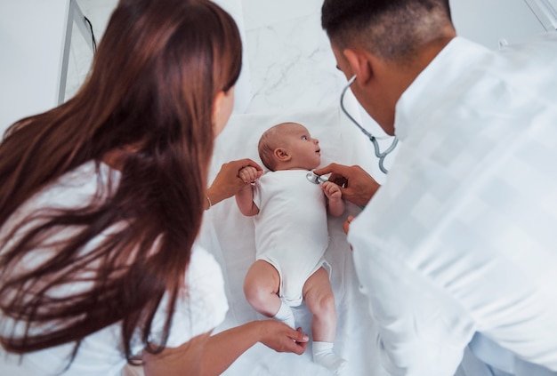 Listening by using stethoscope. Young pediatrician is with little baby in the clinic at daytime.