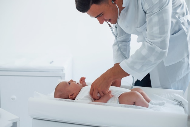 청진기를 사용하여 듣기. 젊은 소아과 의사는 낮에 병원에 작은 아기와 함께 있습니다.