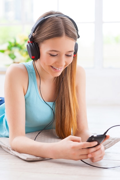 Listening to her favorite music. Cheerful teenage girl in headphones listening to the music while lying down on the floor at her apartment