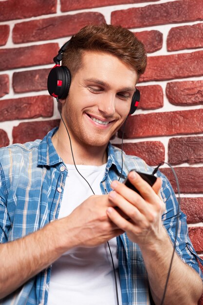 Listening to his favorite music. Handsome young man listening to the MP3 Player while standing against brick wall