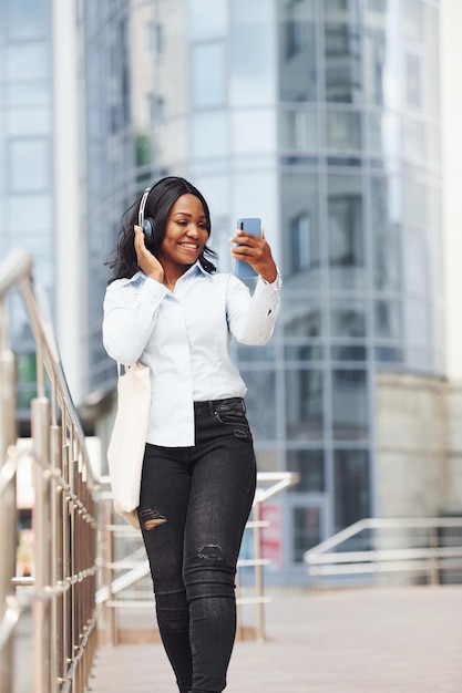 Listening to music in headphones Young afro american woman in white shirt outdoors in the city against business building