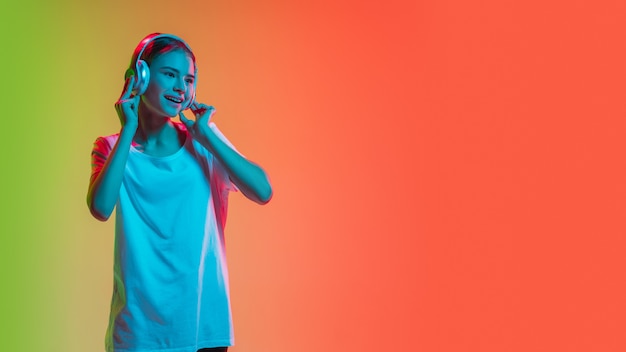 Listening to music with headphones. Young caucasian girl's portrait on gradient green-orange studio background in neon light. Concept of youth, human emotions, facial expression, sales, ad. Flyer