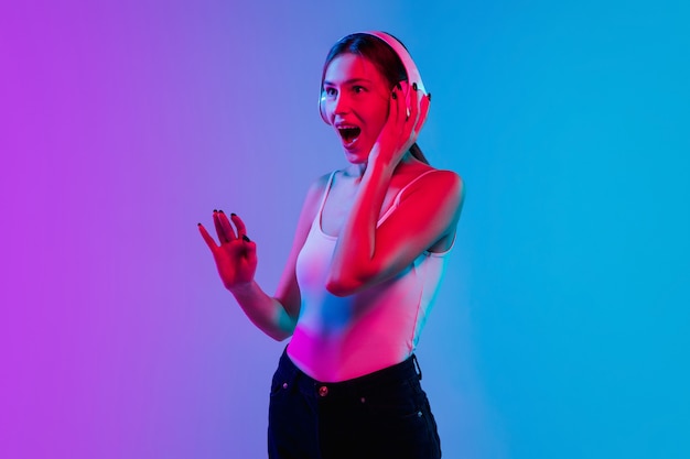 Listening to music with headphones. Young caucasian woman's portrait on gradient blue-purple studio background in neon. Concept of youth, human emotions, facial expression, sales, ad. Beautiful model.