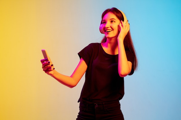 Listening to music with headphones. Young caucasian woman's portrait on gradient blue-yellow studio background in neon. Concept of youth, human emotions, facial expression, sales, ad. Beautiful model.