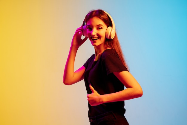 Listening to music with headphones. Young caucasian woman's portrait on gradient blue-yellow studio background in neon light. Concept of youth, human emotions, facial expression, sales, ad.