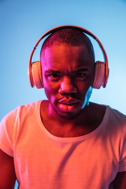 Listening to music Young displeased African man in headphones isolated on blue background in colored neon light Concept of human emotion facial expression youth culture Copy space for ad design