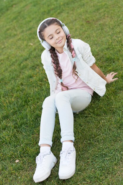 Listening to soft music sound is quite relaxing to me Small girl listening to sound track on green grass Little child wearing earphones playing electronic sound Cute kid enjoying melodious sound