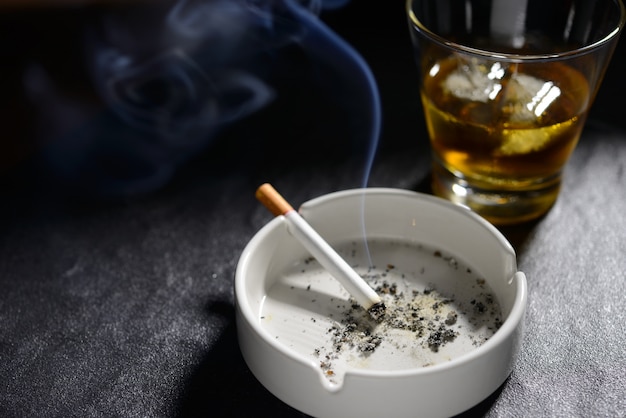 Зажженная и курящая сигарета в пепельнице со стаканом виски