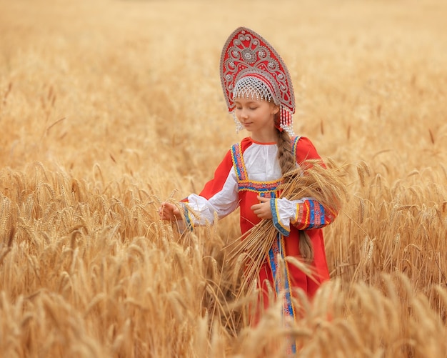 ロシアの国民サラファンのLittlの女の子の子供とsummeの黄金の麦畑に立っているココシニク