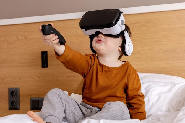 Little adorable boy sitting on bed at home with VR headset and playing interactive video game exploring virtual reality Cute child wearing VR glasses Future gadgets technology education online